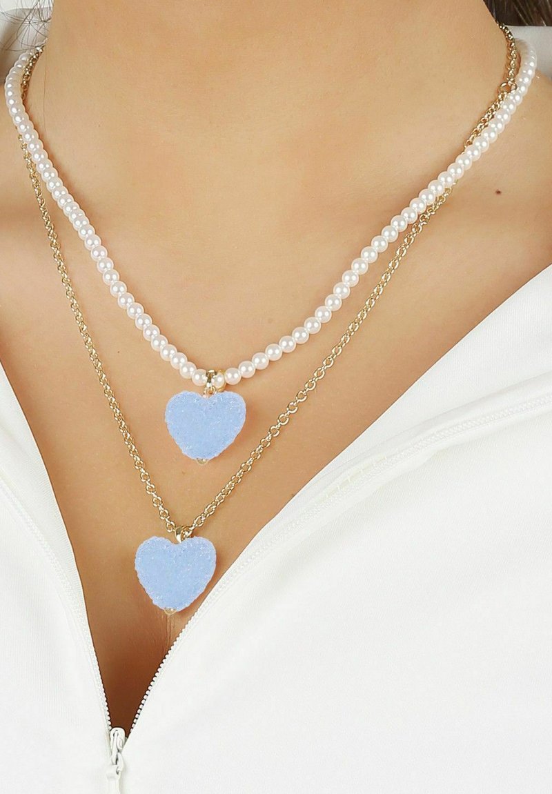 Dubbel lagerhalsband med en rad vita pärlor och en guldkedja, prydd med två blå fluffiga hjärtan som hängsmycken.