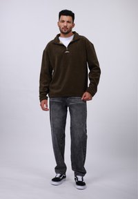 Pull en polaire vert olive foncé avec un col zippé à mi-hauteur et un petit logo. Porté avec un jean gris et des baskets noires à semelles blanches.