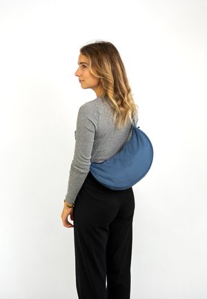 Blå tygaxelväska med en böjd form, slät yta och logotydelse, bärs i crossbody-stil över en grå långärmad tröja.