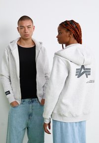 Hoodie gris zippé en matériau doux, avec un logo dans le dos, des poches avant et une capuche avec cordon. Porté avec un jean décontracté.