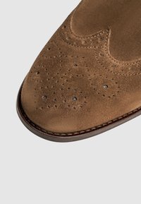 Chaussure en daim marron avec des détails perforés, un capuchon lisse et des coutures contrastantes le long de la semelle.