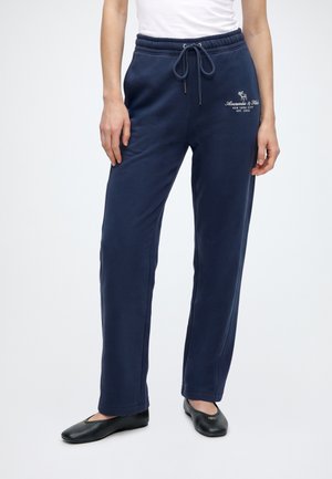 Vrouw draagt marineblauwe joggingbroek met trekkoord en wit logo op de heup, gecombineerd met zwarte ballerina's en een wit topje.