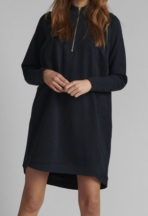 Femme portant une robe sweat-shirt bleu marine à manches longues avec un col à fermeture éclair moitié, debout les mains jointes devant un fond gris uni.
