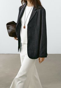 Blazer en denim foncé avec revers crantés, deux boutons et poches avant, associé à un haut blanc côtelé et un pantalon blanc. Accessoire sac tressé marron.