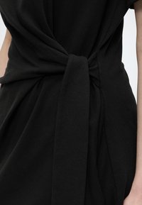 Robe en tissu noir nouée en nœud à la taille, laissant voir une partie des bras du porteur sur un fond clair uni.