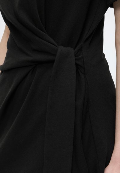 Robe en tissu noir nouée en nœud à la taille, laissant voir une partie des bras du porteur sur un fond clair uni.