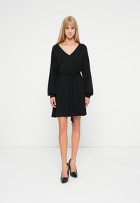 Vestido negro con escote en V y mangas largas abullonadas. Incluye un cinturón atado en la cintura, textura suave y diseño a la altura de las rodillas. Llevado con tacones negros.