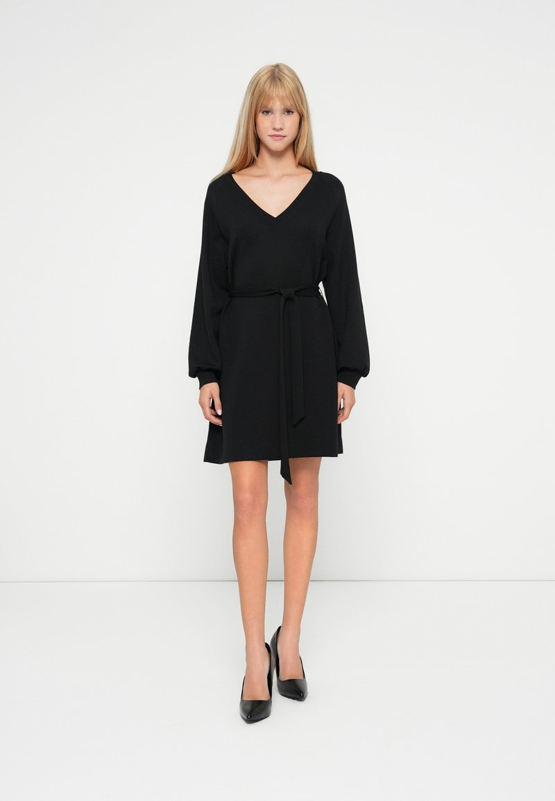 Vestido negro con escote en V y mangas largas abullonadas. Incluye un cinturón atado en la cintura, textura suave y diseño a la altura de las rodillas. Llevado con tacones negros.