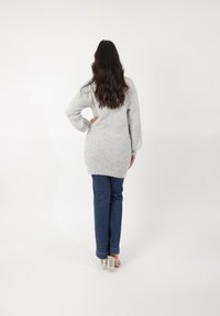 Pull gris surdimensionné avec tricot texturé, associé à un jean skinny bleu foncé. Chaussures à talons transparents. Modèle de dos.