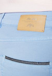 Jean en denim bleu clair avec un patch en cuir embossé d'un texte et une fermeture éclair décorative sur la poche arrière. Texture lisse et coupe ajustée.