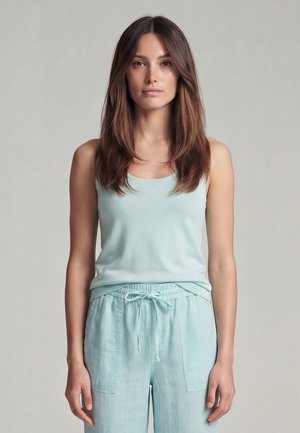 Young woman with long brown hair wearing a light blue sleeveless top and matching drawstring pants, standing against a plain background.