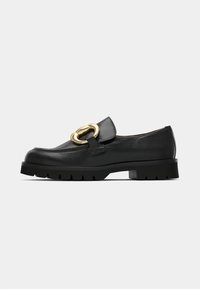 Zwarte leren instap-loafer met dikke zool en grote gouden gesp bovenop, gepresenteerd op een witte achtergrond.