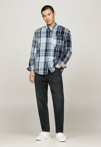 Camisa de cuadros en azul y carbón con un bolsillo en el pecho y detalle de logo, combinada con jeans oscuros y zapatillas blancas.
