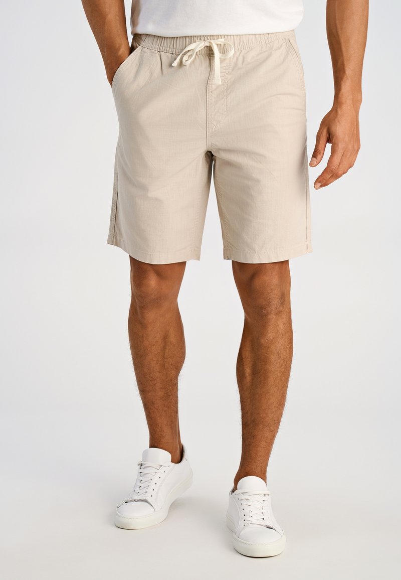 Pantalones cortos beige con cinturilla elástica y cordón, con bolsillos laterales y corte recto. Usados con zapatillas blancas sobre un fondo neutro.