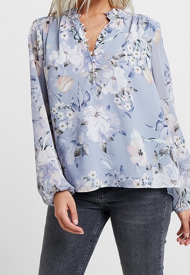 Blouse bleu clair à manches longues avec imprimé floral blanc et violet, col à volants, associée à un jean gris foncé.
