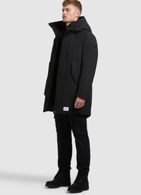 Parka neagră, până la genunchi, cu guler înalt, buzunare frontale și o etichetă cu logo la tiv. Purtați cu pantaloni cargo negri și cizme.