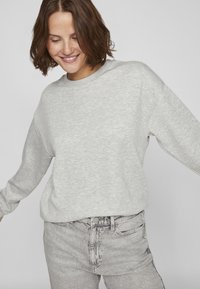 Mujer sonriente con cabello corto castaño, vistiendo una sudadera gris clara y jeans de tiro alto de un gris desgastado, contra un fondo plano.