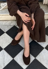 Robe marron ample en tissu texturé avec des manches bouffantes, associée à des chaussures mocassins en daim marron foncé, sur un sol carrelé noir et blanc.