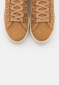 Zapatillas de corduroy marrón con parte superior texturizada, que presentan ojales en oro rosa y cordones planos beige, sobre un fondo blanco.