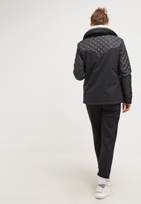 Veste matelassée noire avec un corps noir lisse contrastant, col doublé en polaire, et pantalon noir assorti, accompagnés de baskets de couleur claire.