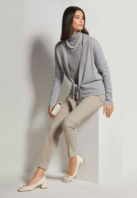 Grijze cardigan met strikdetail over een grijze coltrui, beige slim fit broek en crèmekleurige block-heeled schoenen, met een kleine clutch tas.