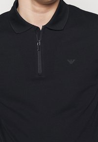 Černá polo tričko na zip s límečkem, s jemně vyšívaným logem na hrudi a hladkým, texturovaným materiálem.