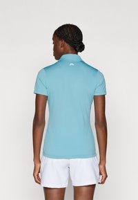 Světle modrá polo tričko s krátkým rukávem a límcem, hladký materiál a malé logo na zadní straně; v kombinaci s bílými šortkami.