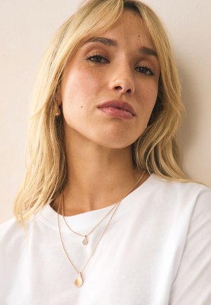 Blonde woman wearing white shirt with two layered gold necklaces featuring teardrop-shaped pendants, neutral background.
