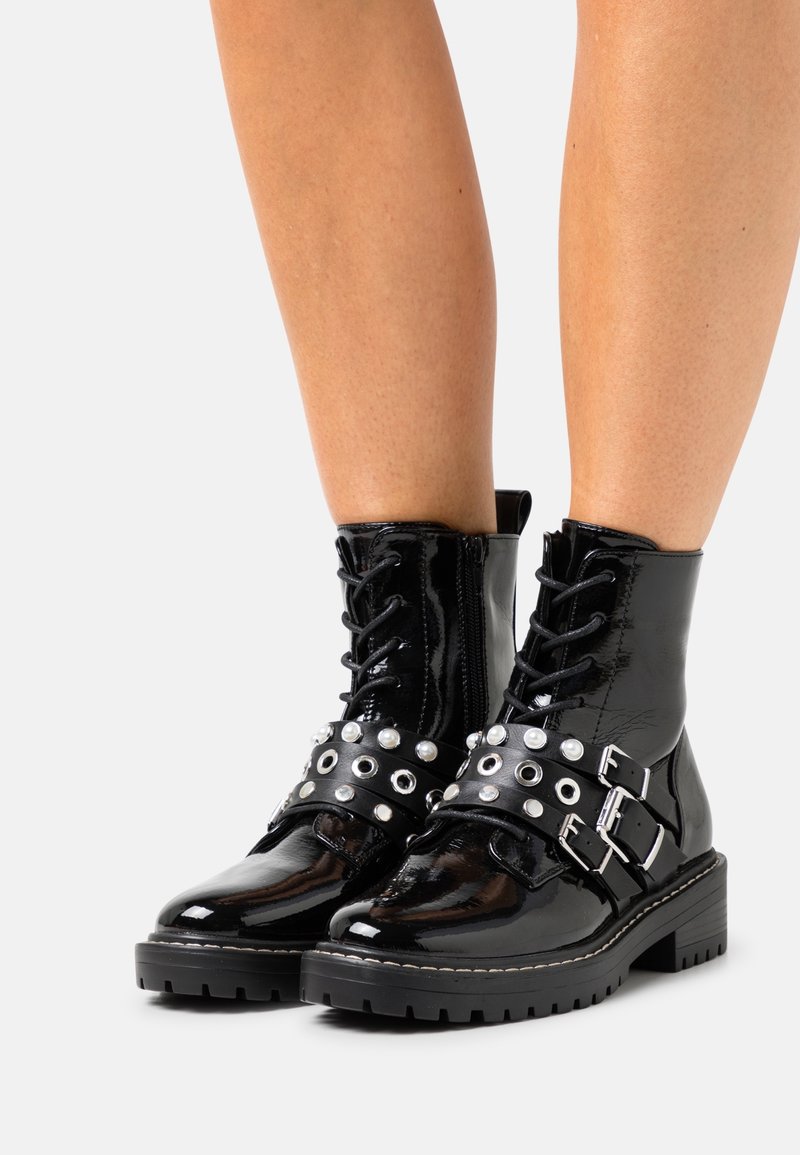 Bottines en cuir verni noir avec fermeture à lacets sur le devant, brides cloutées, détails en métal argenté et semelle épaisse.