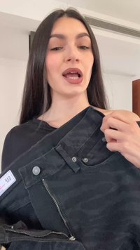 Mlada ženska z dolgimi temnimi lasmi, ki drži in kaže na odprte črne hlače v notranjih prostorih.