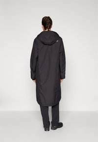 Langer schwarzer Mantel mit Kapuze, glatte Textur und gerade Silhouette, getragen über dunklen Hosen; einfaches und funktionales Design.
