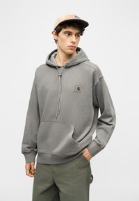 Νέος άνδρας με γκρι φούτερ Carhartt και παντελόνι ελιάς, φοράει μπεζ καπέλο, στέκεται με το ένα χέρι στην τσέπη και κοιτάζει προς τα πλάγια.