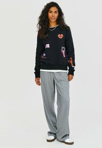 Čierna mikina s farebnými nášivkami, dlhé rukávy, hrubé rebrované manžety, v kombinácii so šedými širokými nohavicami s prúžkami a bielymi plochými šľapkami.