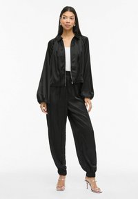 Černá saténová bunda na zip s nashromážděnými rukávy, spojená s černými saténovými kalhotami s elastickým pasem. Modelka má na sobě bílé tričko a stříbrné podpatky.