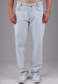 Mann in hellblauen Jeans und weißen Sneakers, der mit entspannten Händen an den Seiten vor einem schlichten grauen Hintergrund steht.