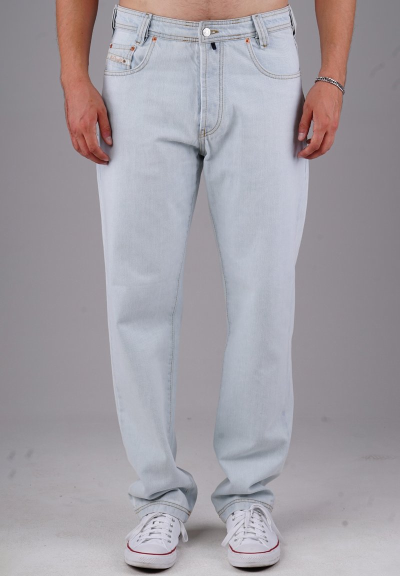 Mann in hellblauen Jeans und weißen Sneakers, der mit entspannten Händen an den Seiten vor einem schlichten grauen Hintergrund steht.