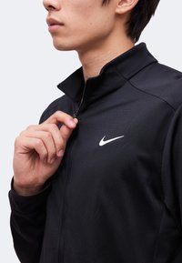 Person trägt eine schwarze Nike-Jacke, hält den Reißverschluss nahe am Kragen, mit dem weißen Nike-Logo sichtbar auf der Brust.