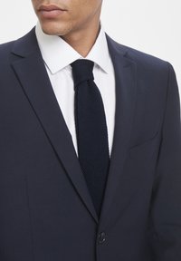 Blazer azul marino con solapas de muesca, cierre de botones y un solo bolsillo en el pecho, combinado con una camisa blanca y una corbata de punto azul oscuro texturizada.