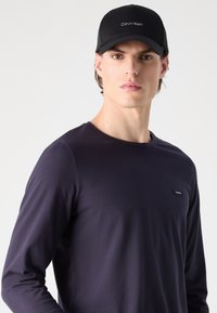 Μαύρο καπέλο μπέιζμπολ με λογότυπο "Calvin Klein", συνδυασμένο με εφαρμοστό ναυτικό μπλουζάκι μακρυμάνικο που διαθέτει μικρή πλάκα λογότυπου στο στήθος.