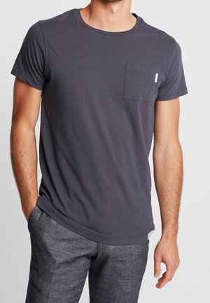 Mężczyzna ubrany w gładki ciemnoszary t-shirt z krótkim rękawem i kieszenią na piersi oraz szare spodnie o fakturze, stoi z jedną ręką w kieszeni.