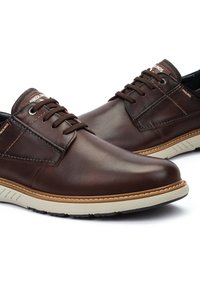 Zapatos de cuero marrón con un acabado liso, que cuentan con un diseño de cordones, suela de goma blanca con un acento marrón y detalles de marca en el lateral.