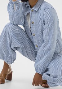 Pruhovaný overal z modré a bílé látky s límečkem, zapínáním na knoflíky, elastickými manžetami a volně střiženými kalhotami. Hnědé sandály na podpatku doplňují tento outfit.