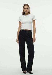 Bílý přiléhavý krátký rukáv crop top v kombinaci s černými kalhotami s vysokým pasem a širokými nohavicemi. Oblečení doplňuje stříbrný výrazný náhrdelník.