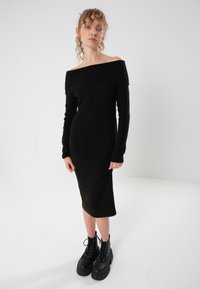 Svart ribbestrikket off-shoulder kjole med lange ermer, tettsittende silhuett, til midt på leggen, kombinert med kraftige svarte snørestøvler.