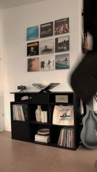 Černá regálová jednotka s různými přihrádkami, zobrazující vinylové desky, dekorativní vázu, knihy, reproduktor a hodiny.