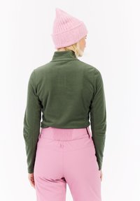 Grøn fleece trøje med høj krave og lange ærmer, parret med lyserøde bukser med en struktureret talje. Uld lyserød hue på hovedet.