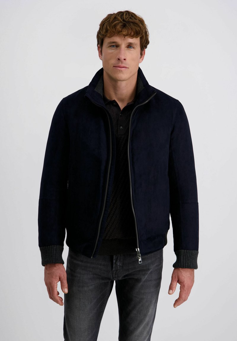 Marineblauwe suède jas met een hoge kraag, voorrits en geribde gebreide manchetten. gedragen over een zwarte trui, gecombineerd met donkere jeans.