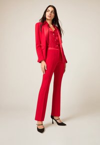 Rood op maat gemaakt pak met een aansluitende blazer en bijpassende broek, gemaakt van gladde stof, met een subtiele glans en een strakke silhouet. Zwarte hakken.