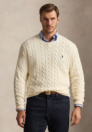 Homme portant un pull en maille torsadée crème par-dessus une chemise bleu clair à col, jean foncé et ceinture marron, sur fond uni.