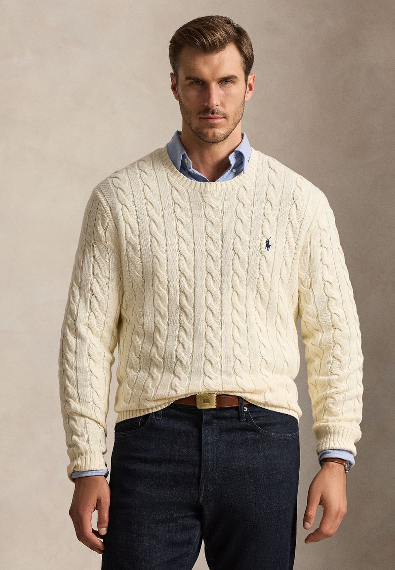 Uomo che indossa maglione crema a trecce sopra camicia azzurra con colletto, jeans scuri e cintura marrone su sfondo semplice.